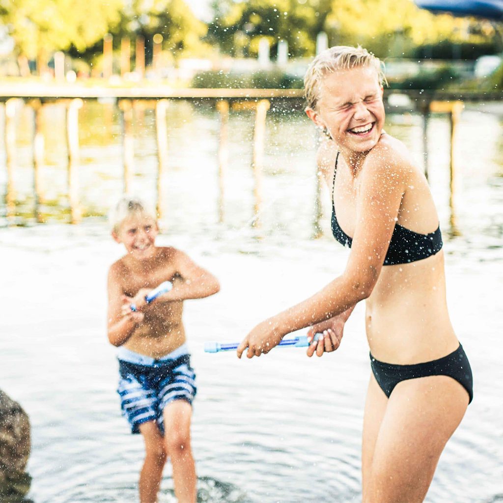 TOURISMUSFILM 47 Zwei Kinder spielen im Wasser eines Strandbads mit Wasserpistolen, Prienavera Erlebnisbad & Strandbad, Lifestylefotografie, WINGMEN Media